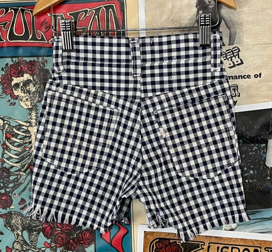 Vintage 1970s kids navy blue and white gingham plaid white tab Levi's cutoff shorts displayed on a hanger against a colorful backdrop featuring vintage advertisements.