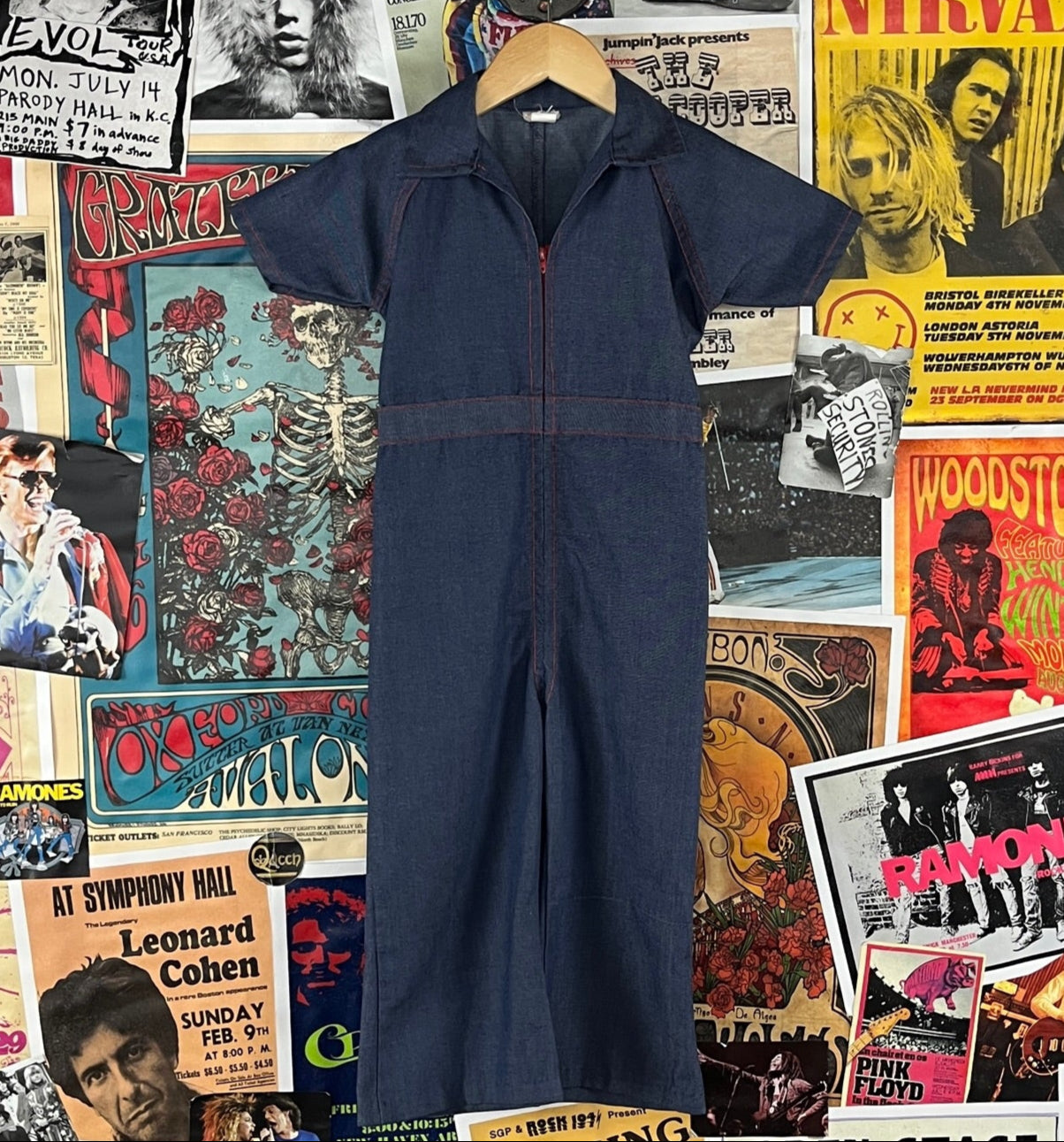 Vintage toddler kids boys or girls retro 1970s blue short sleeve collared flared zip up denim jean jumpsuit by JCPenney. Features red stitching and a red zipper, as well as an elastic back waist. Made of 50% polyester and 50% cotton. Has a minor manufacturing error on the leg, but are in otherwise good, pre-owned condition. Are tagged a kids size 4.