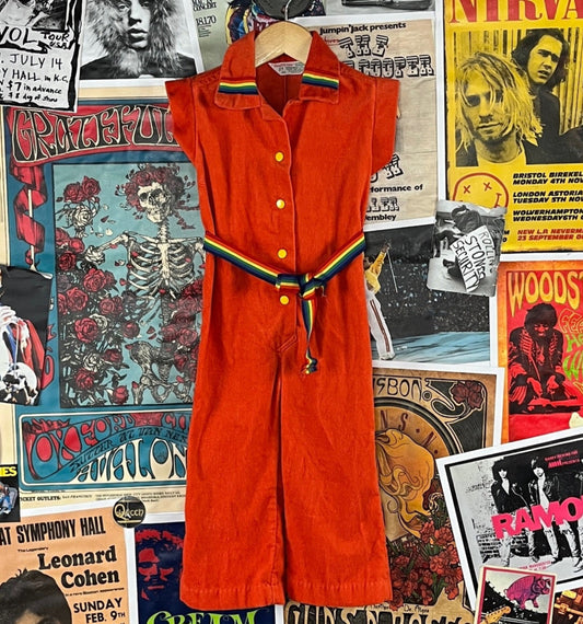 Awesome vintage toddler kids retro 1970s orange corduroy short cap sleeve jumpsuit with rainbow trim collar, rainbow belt, and elastic back waist by Health-tex. Features yellow snap buttons up the front. Is made of 50% polyester and 50% cotton in the U.S.A. Tagged size 2T. We estimate it to best fit size 18-24 months. Has a light spot on the front left chest, but is in otherwise great, pre-owned condition.
